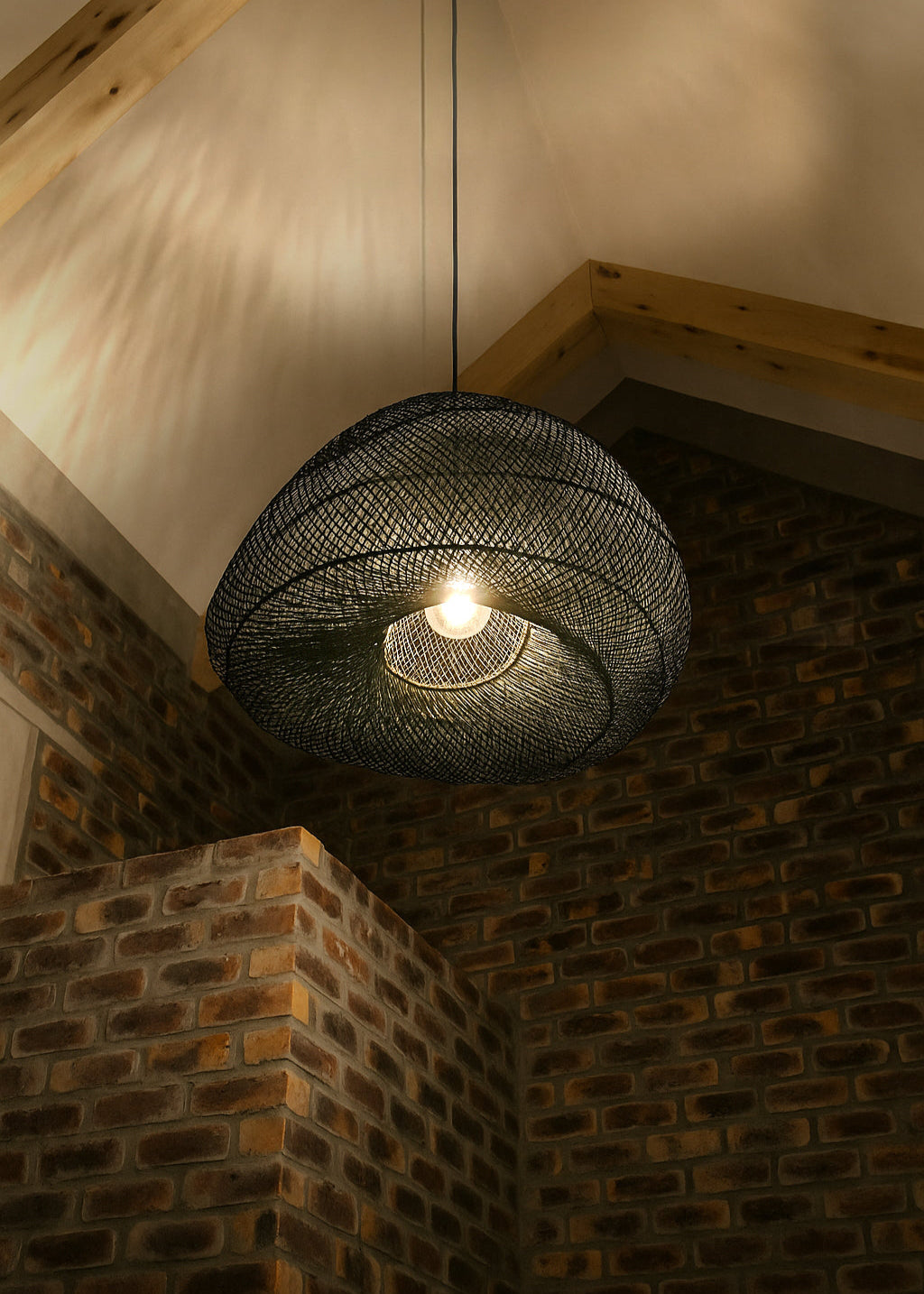 Woven black pendant light hanging from a wooden ceiling with a brick wall background