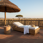 Wicker lounge chair with cushions under a thatched umbrella on a sunlit deck with a scenic view.