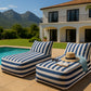 A rectangular daybed lounger with a bold blue and white striped fabric, featuring a low, boxy base and a matching angled backrest cushion. The clean white background highlights the modern, casual design and crisp piping detail along the seams.
