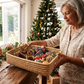An older woman with gray hair holds a woven rectangular  Malawi Furniture basket filled with colorful Christmas ornaments and pinecones in a warmly lit indoor setting with a decorated Christmas tree in the background.