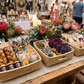 A rustic market stall display featuring Malawi Furniture woven baskets filled with artisanal treats, including twisted brown pastries, jars of spread, and colorful giraffe-shaped chocolates, alongside patterned drawstring pouches, set against a blurred background of flowers and shoppers.