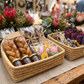 A rustic market stall display featuring Malawi Furniture woven baskets filled with artisanal treats, including twisted brown pastries, jars of spread, and colorful giraffe-shaped chocolates, alongside patterned drawstring pouches, set against a blurred background of flowers and shoppers.