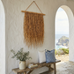 Hanging Grass Wall Decor in a coastal villa. Handcrafted hanging grass wall art by Malawi Furniture, featuring natural dried organic ilala palm and seagrass onto a sustainable wooden dowel. Large 90cm by 1m boho fringed wall hanging in a light natural brown tone for minimalist home decor.